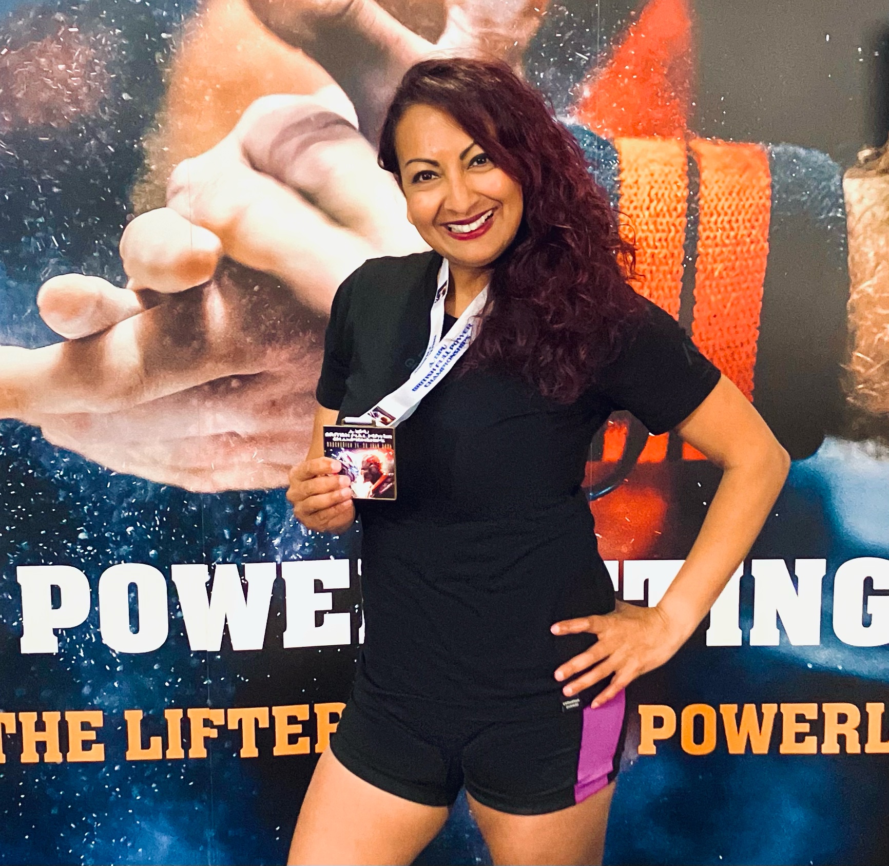 Aneela Rose British Championships Portrait Aneela Rose holding a medal in front of a powerlifting mural, alternate view 2
