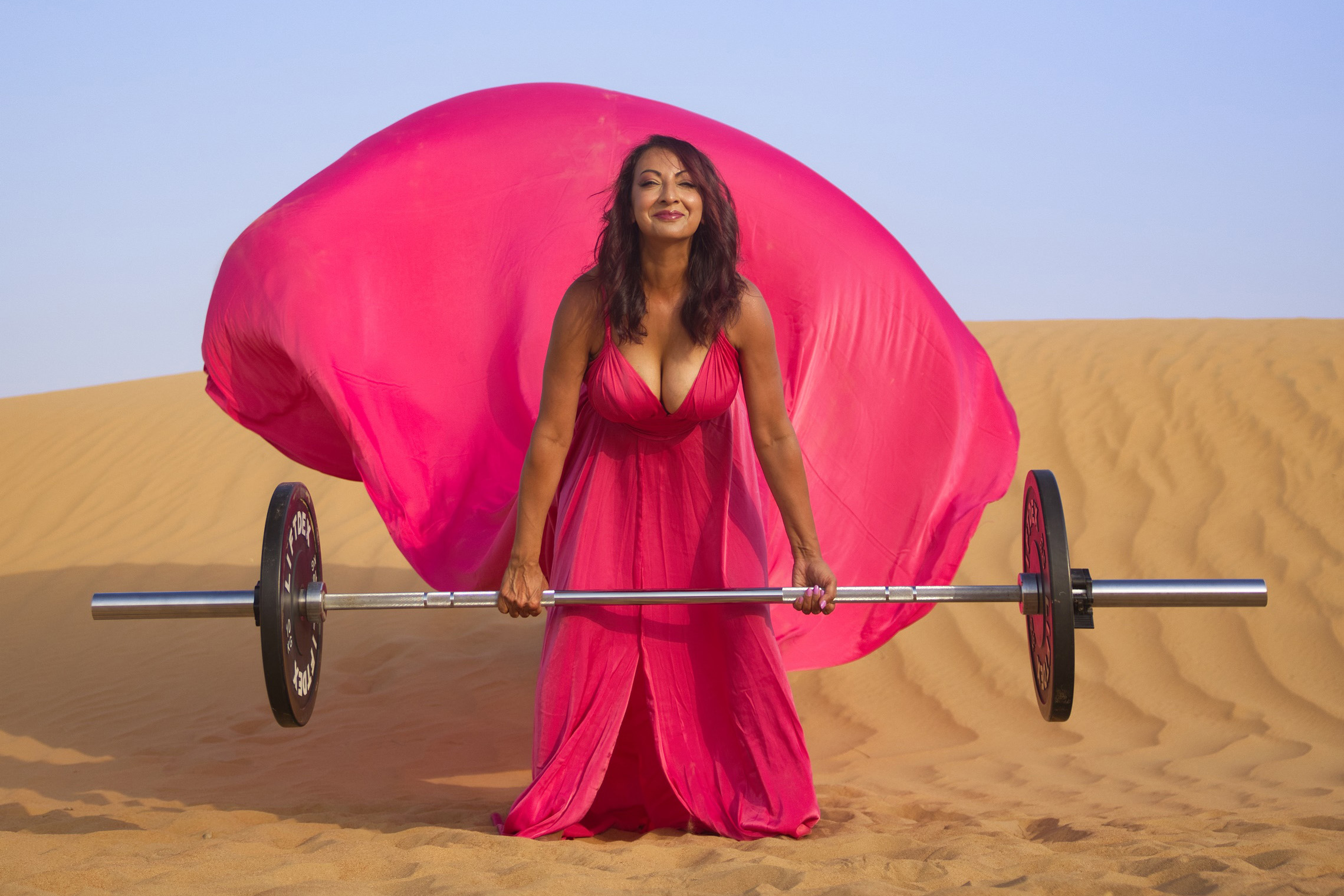 Aneela Rose Pink Dress Desert Deadlift Wide Wide view of Aneela Rose crouched behind a barbell in a flowing hot pink dress in the desert