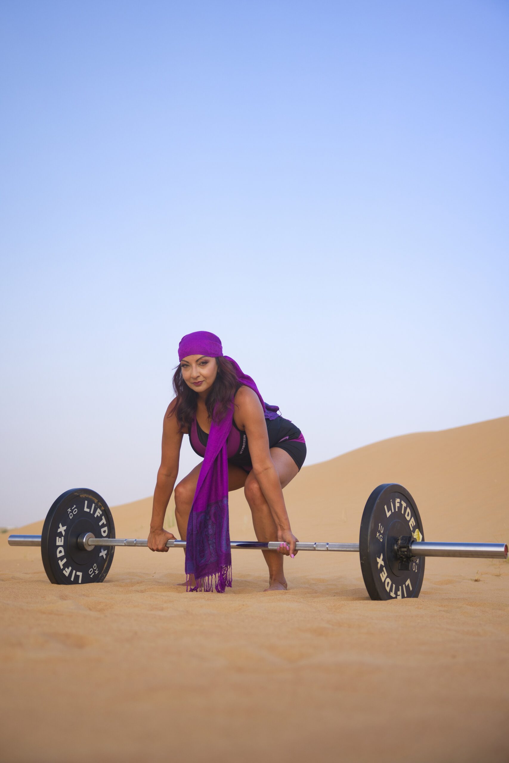 Aneela Rose Crouched by Desert Barbell Aneela Rose crouched beside a barbell in the desert wearing a purple headscarf