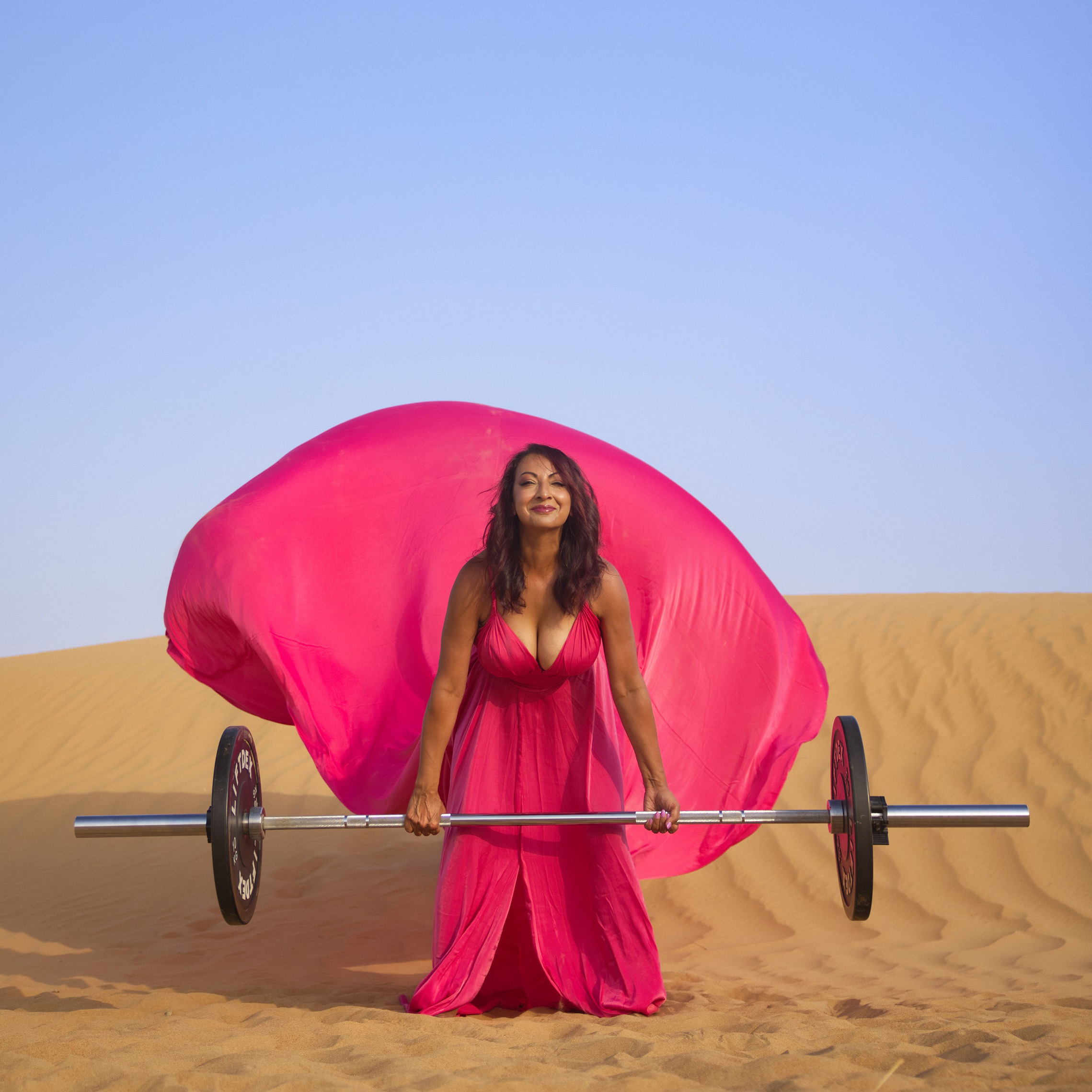 Aneela Rose Desert Deadlift in Pink Dress Aneela Rose crouched behind a barbell in a flowing hot pink dress in the desert