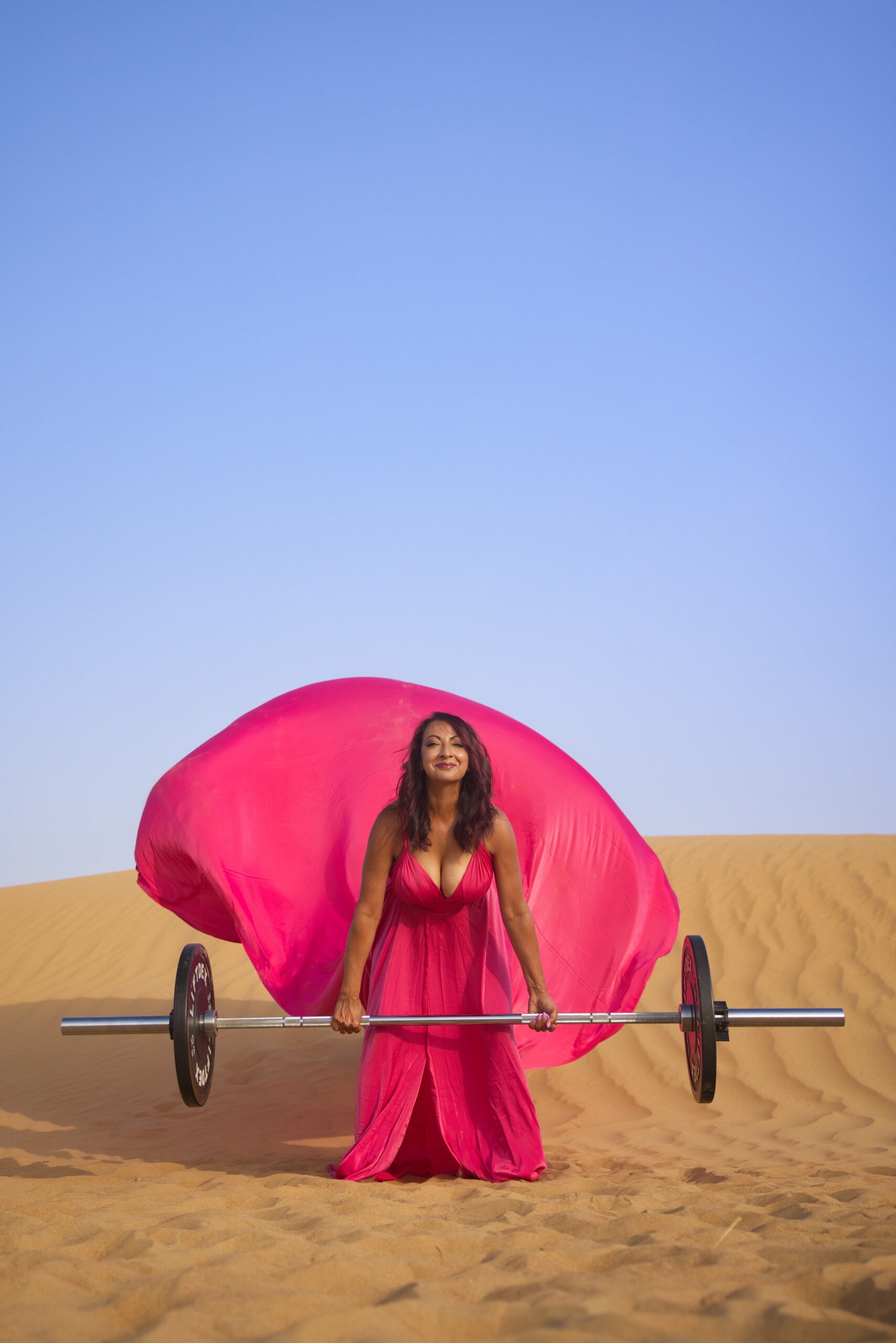 Aneela Rose Pink Dress Desert Deadlift Aneela Rose crouched holding a barbell while wearing a flowing hot pink dress in the desert